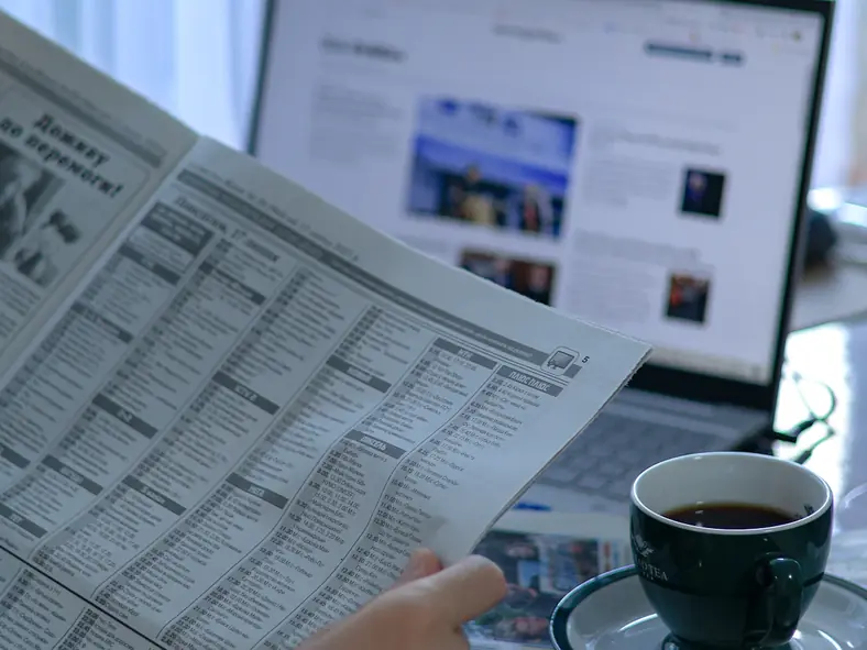 Reader holding a newspaper next to a laptop showing a news website